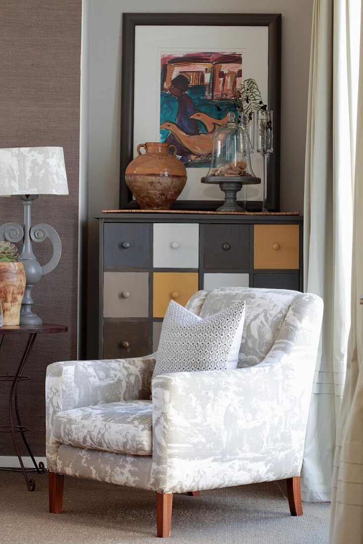 White, lustrous armchair in front of chest of drawers with multicoloured drawers in niche