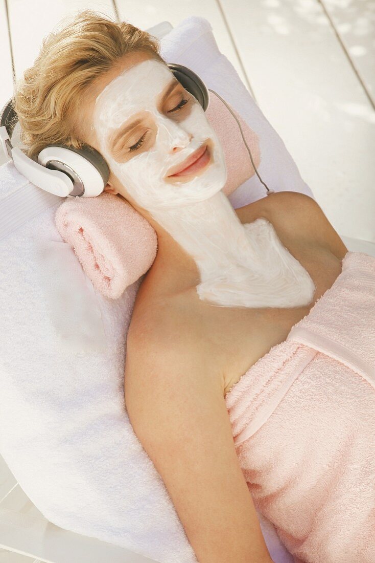 A blonde woman wearing headphones relaxing with a nourishing mask on her face, neck and chest