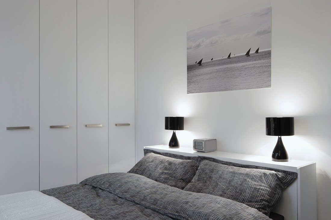 Doppelbett mit grauer, gemusterter Bettwäsche, an Kopfende Ablage mit schwarzen Tischleuchten in modernem Schlafzimmer mit weißem Einbauschrank