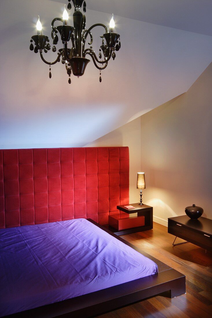 Low, dark wooden bed with purple sheets, red upholstered wall panel and chandelier in small attic room
