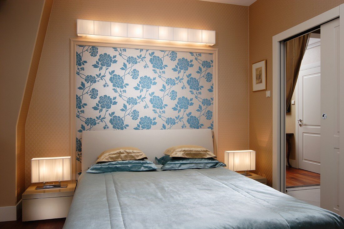 Simple double bed with upholstered headboard and stacked pillows against wall with framed section of floral wallpaper under wall-mounted lamp: open door with view into corridor beyond