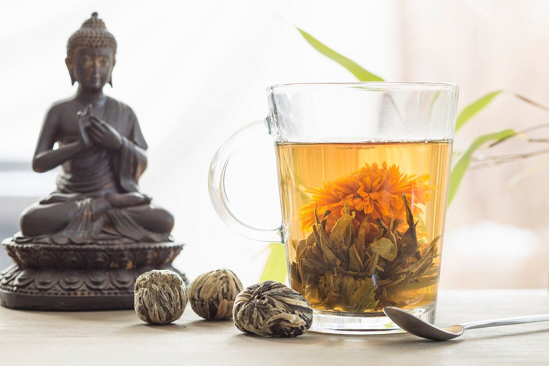 Blütentee im Glas, geschlossene Teeblüten und Buddha im Hintergrund