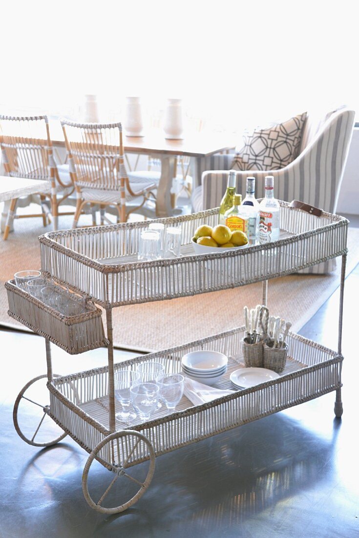 Crockery on delicate serving trolley with lattice trays; wicker chairs and armchair in brightly lit dining area in background