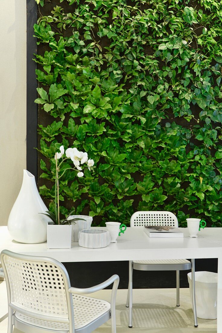 Green wall decorated with faux leaves behind white dining table and woven chairs