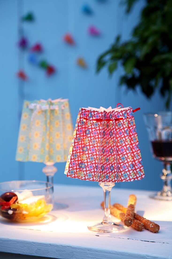 Brightly printed paper folded into concertinas as lampshades for tealights in stemmed glasses