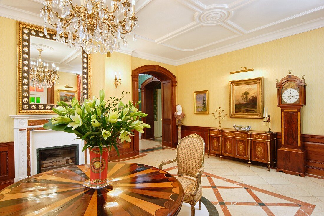 Lilienstrauss auf rundem Holztisch mit Intarsien, unter Kronleuchter in luxuriösem Esszimmer