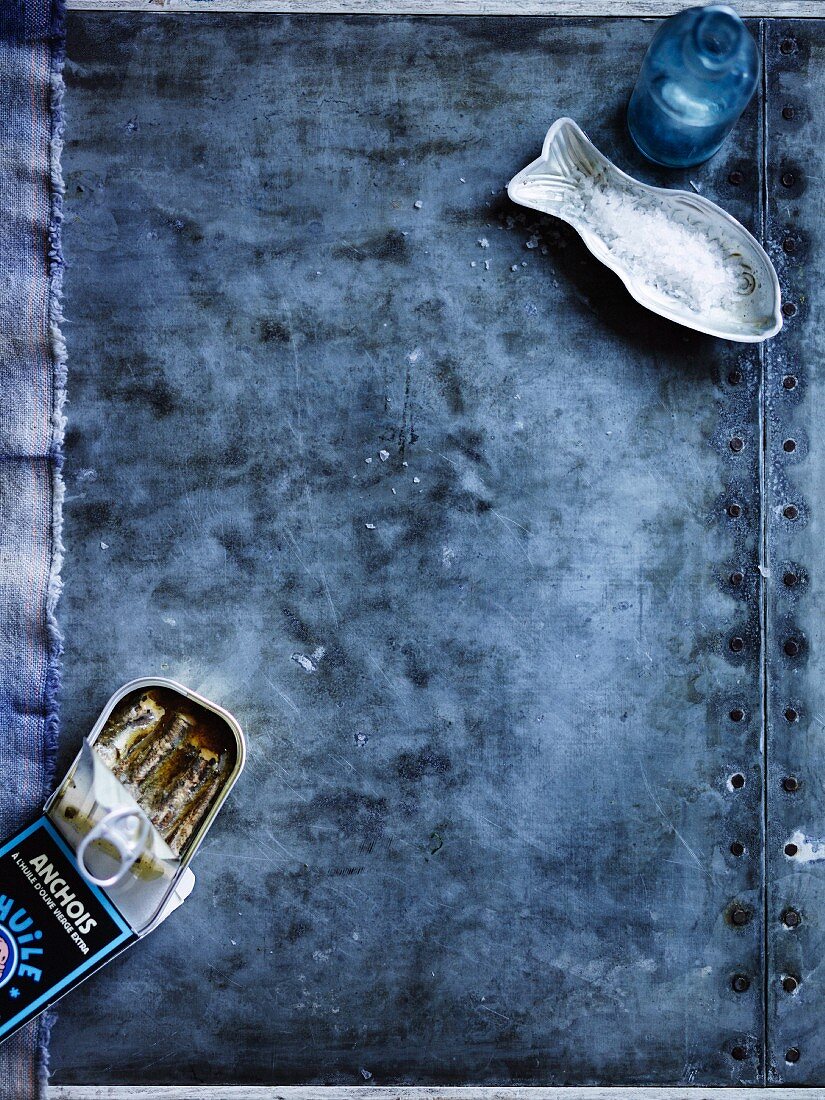 A tin of anchovies and a fish shape with salt on a blue fabric background