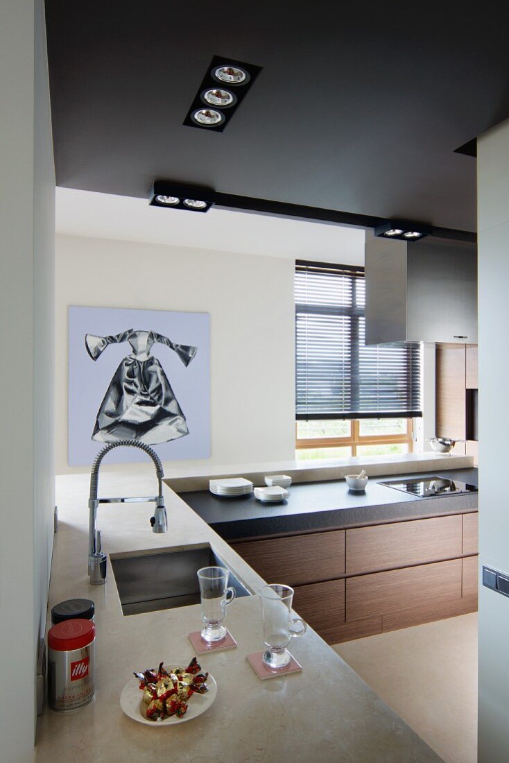 View from open corner into designer kitchen with sink and hob integrated into L-shaped worksurfaces and modern artwork in background