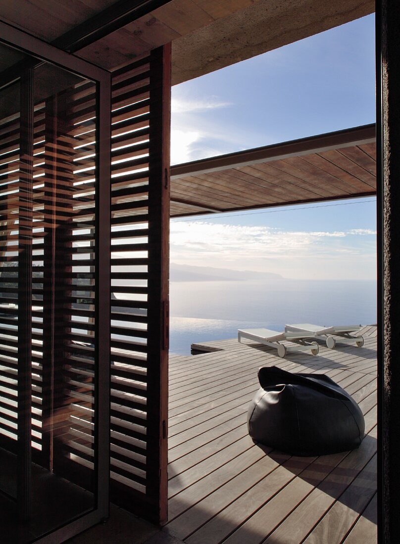 Blick durch offene Lamellentür auf Holzdeck mit Sitzsack und Sonnenliegen vor Meer