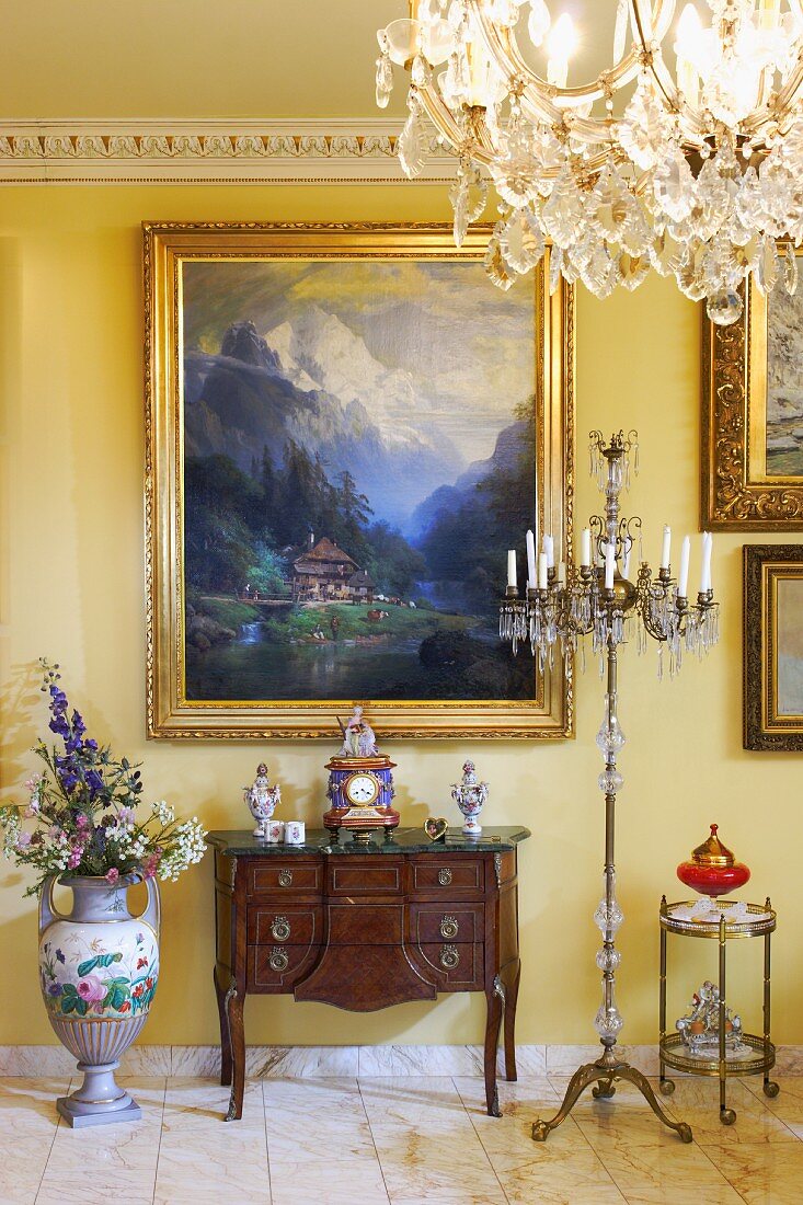 Antique, dark wood bureau below gilt-framed landscape on yellow-painted wall with chandelier floor lamp to one side