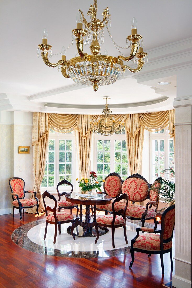 Gilt chandelier in front of period lounge area in semicircular window bay; chairs with red and gold ornamental pattern on upholstery in grand interior
