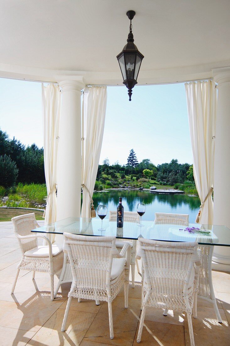 weiße Rattanstühle um Glastisch auf überdachter Terrasse, zwischen Säulen Blick in Garten mit Teich