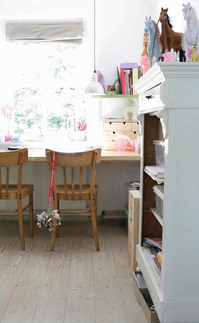 Simple wooden chairs at desk below window and horse ornaments on open-fronted, white-painted cabinet in children's bedroom