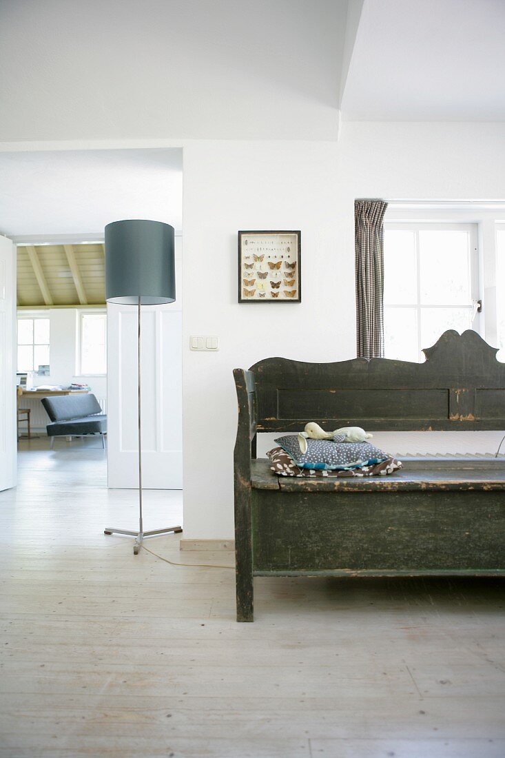 Rustic wooden bench with carved backrest below window next to modern standard lamp with grey lampshade