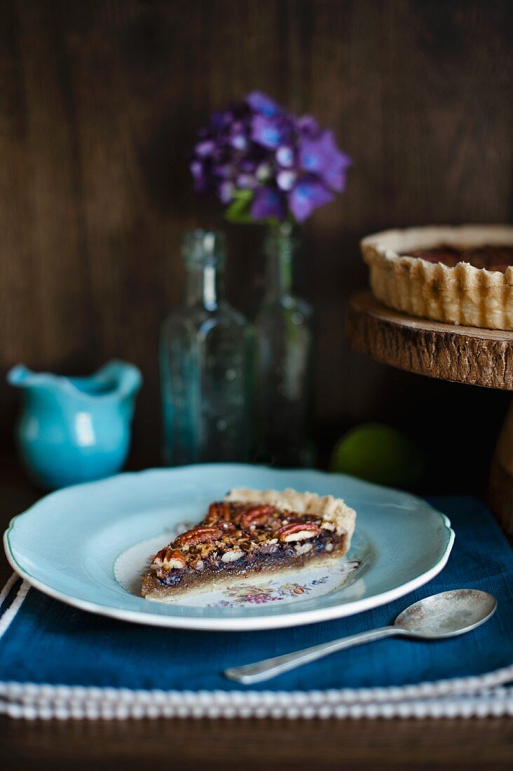 Ein Stück Pecanusstarte auf hellblauem Teller vor Sahnekrug, Blumen und Kuchenständer