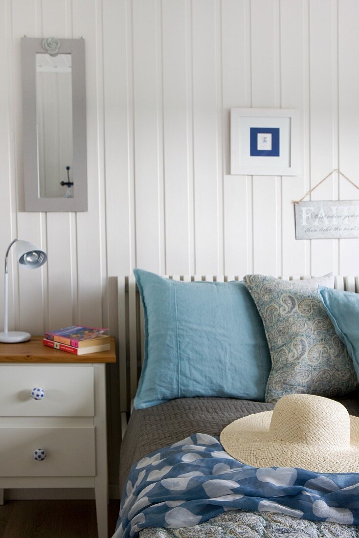 Large pillows and straw hat on bed next to white-painted bedside cabinet against white wooden wall