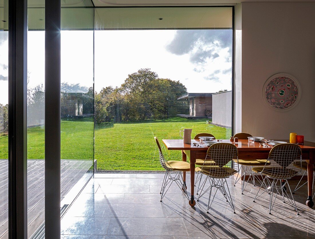 Classic wire-framed chairs around wooden dining table with place settings in front of glass wall with garden view