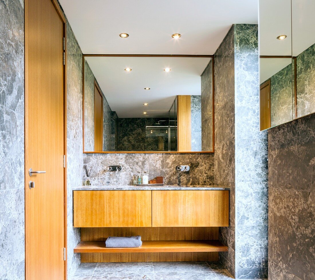 Designerbad mit hellgrau weissen Marmorfliesen an Wänden und Boden, eingebauter Waschtisch mit Unterschrank aus Holz, an Wand vollflächiger Spiegel