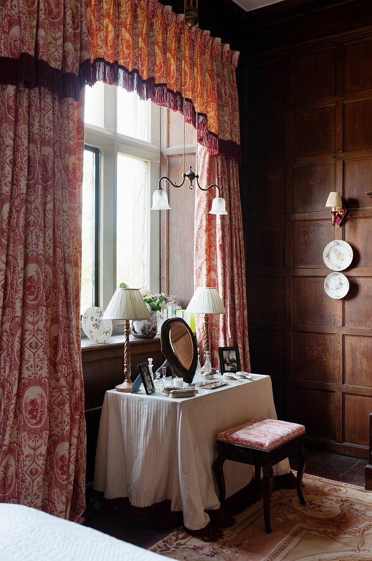 Dressing table below window with toile-de-jouy curtains and decorative china wall plates on wood-panelled walls in bedroom