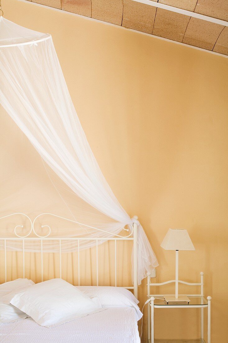 Bett mit filigranem, weißem Metallgestell und luftigem Baldachin in Schlafzimmer mit pastellgelber Wand