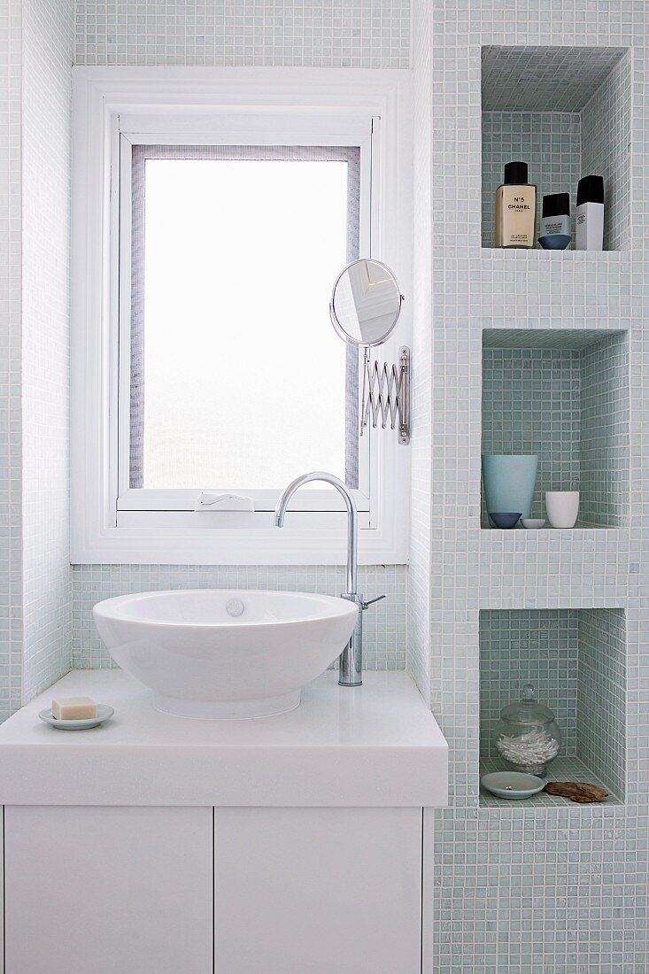 Modern washstand below window and fitted shelf niches with mosaic tiles in bright bathroom
