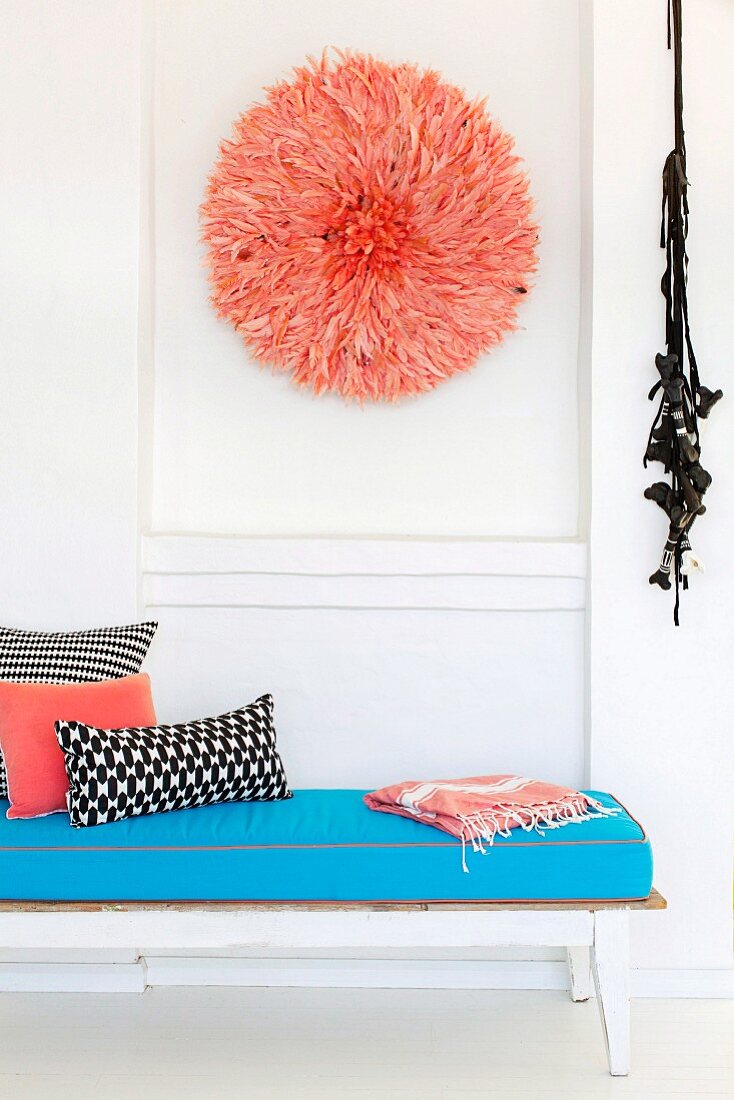 White wooden lounger with turquoise blue upholstery against a white wall with a round feather art object