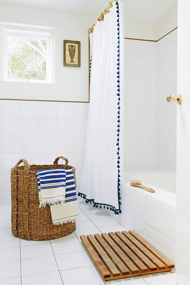Rustikaler Wäschekorb mit blau-weiß gestreiften Handtüchern auf Fliesenboden vor Badewanne mit weißem Duschvorhang