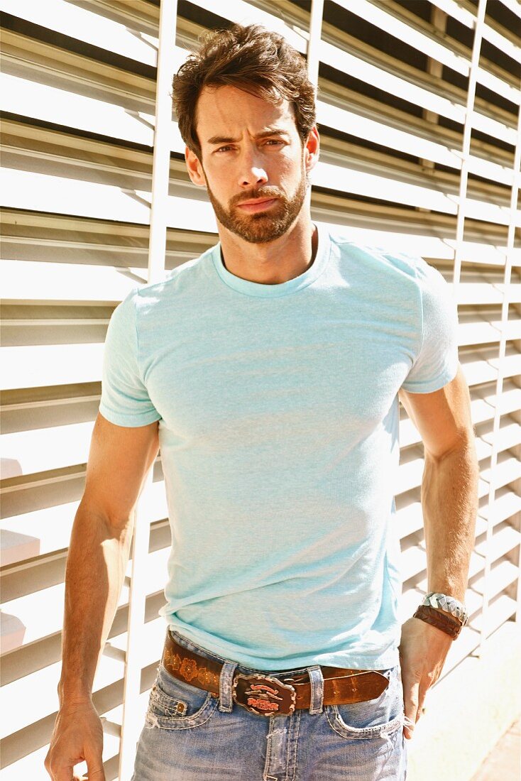 A young man standing in front of white wooden slats wearing a light T-shirt and jeans