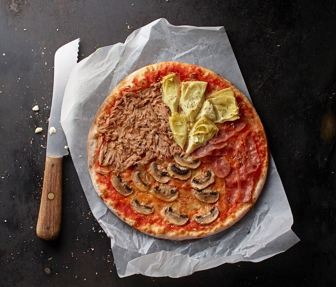 Pizza mit Thunfisch, Champignons, Parmaschinken und Artischocken auf Papier mit Messer