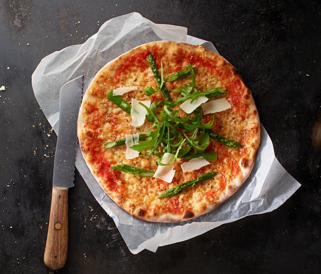 Pizza Margherita mit Spargel, Rucola und gehobeltem Parmesan auf Papier mit Messer
