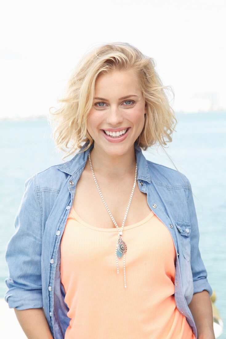 A blonde woman by the sea wearing and apricots top and a denim shirt