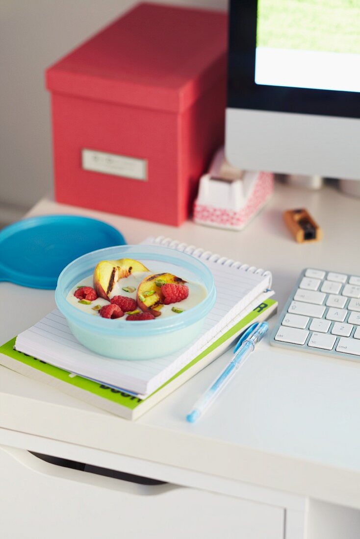 Tupperdose mit Joghurt mit Himbeeren, Grilläpfeln und Pistazien auf Schreibtisch