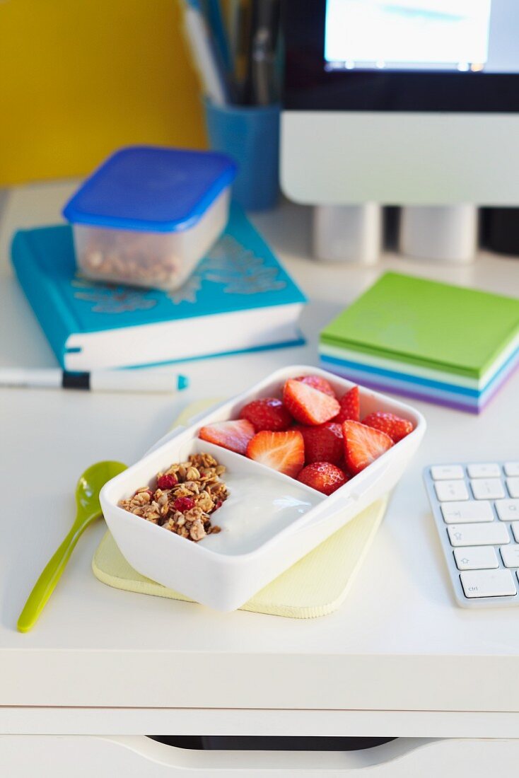Tupperdose mit Joghurtmüsli und Erdbeeren auf Schreibtisch