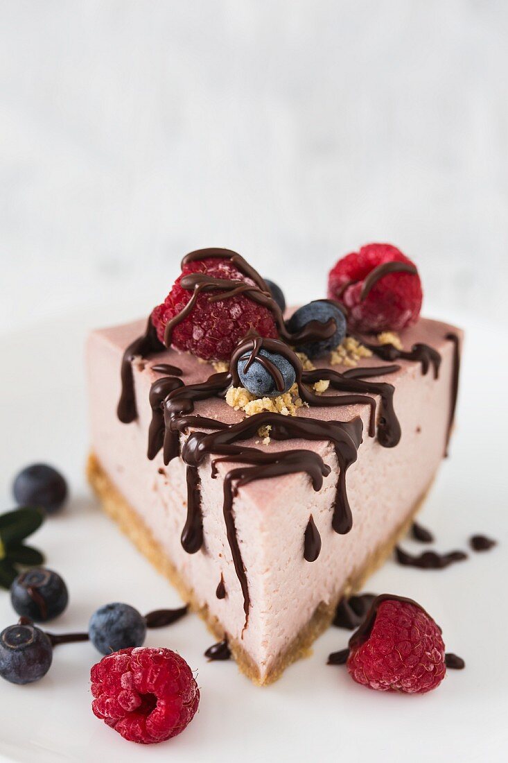 A slice of raspberry cheesecake with fresh raspberries, blueberries and chocolate drizzle