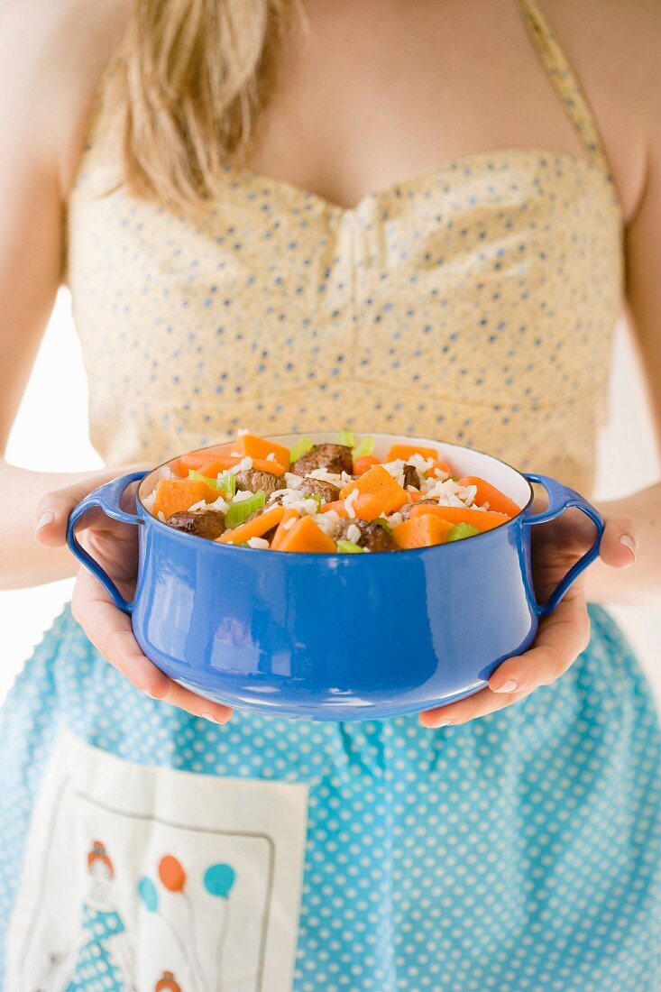 A woman wearing a dress and an apron serving a beef stew with carrots, celery and rice