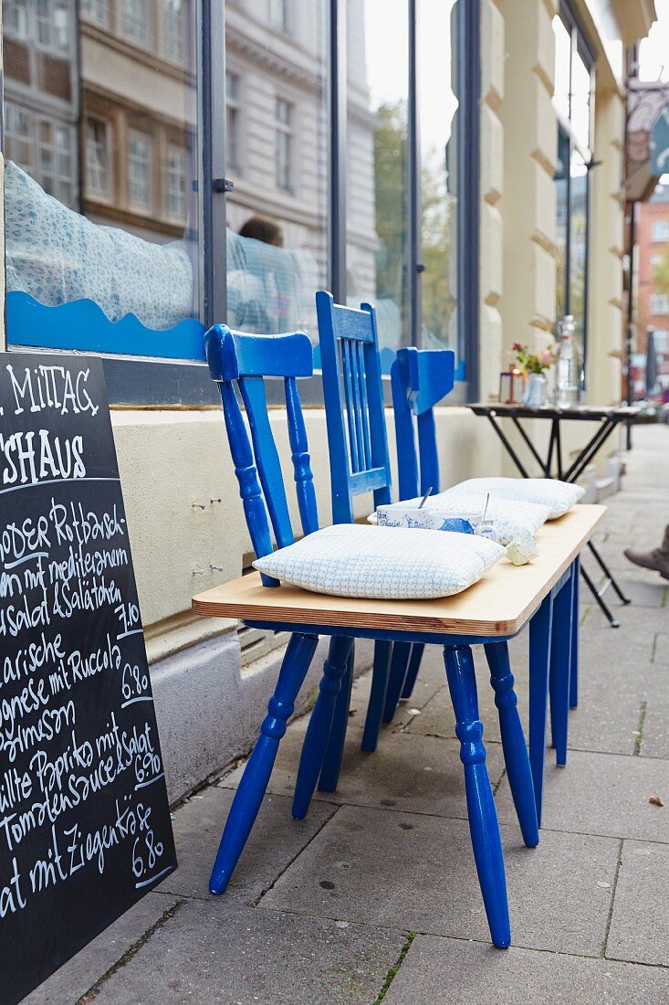 Drei blaue Holzstühle mit Holzplatte zu einer Sitzbank umfunktioniert vor Restaurant