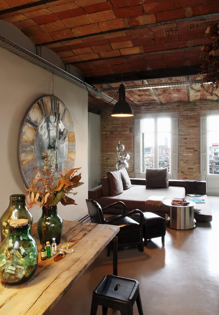 Group of vases on wooden table, 50s-style metal stool and living and sleeping areas in background in rustic interior with brick wall and ceiling