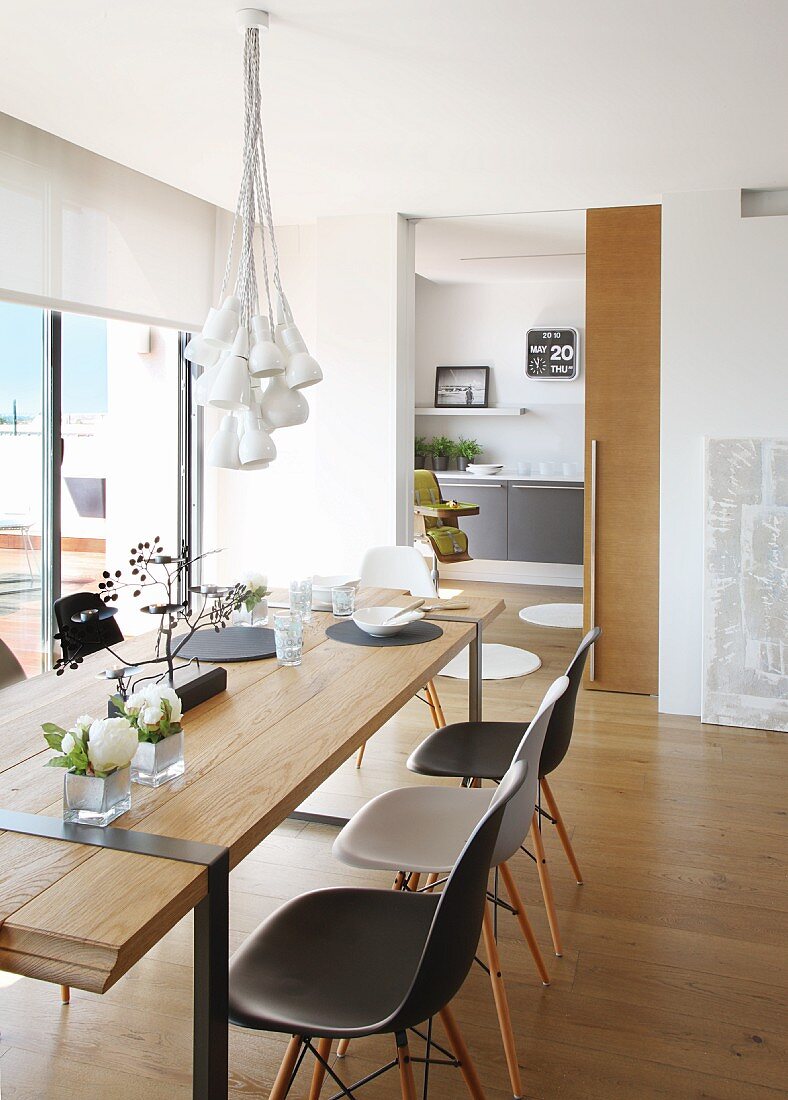 Bundle of lamps above simple, designer wooden table and Eames Plastic Chairs; sliding door leading to kitchen in background