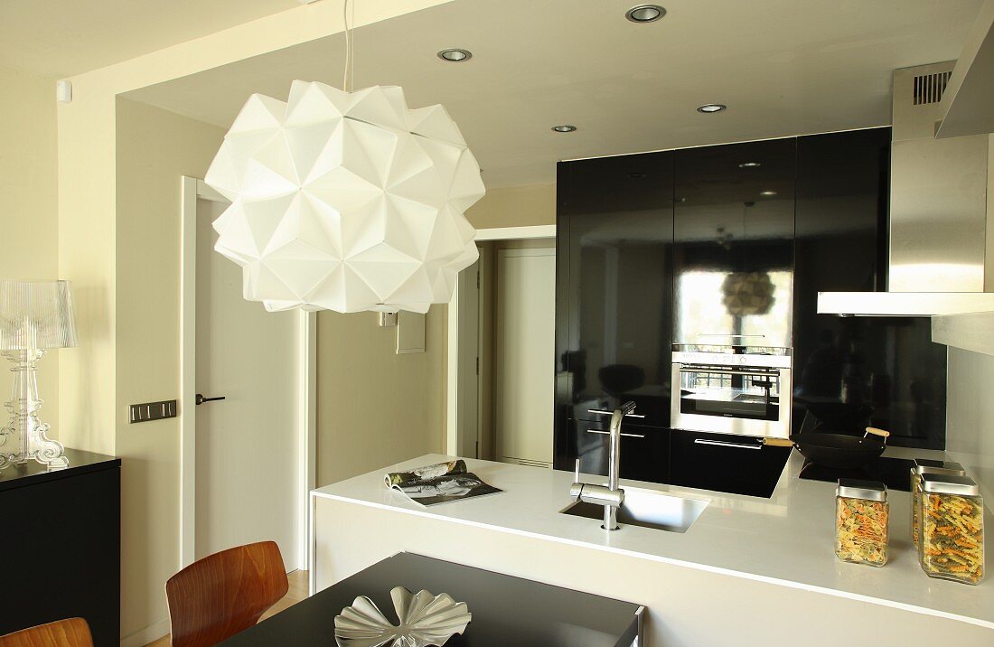 Designer pendant lamp above dining area in front of open-plan kitchen; black, glossy fitted cabinets with appliances in background