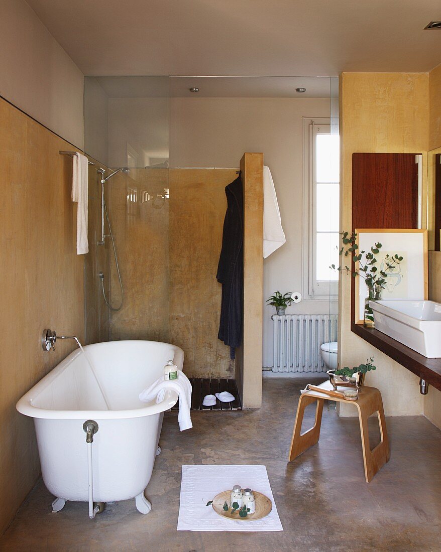 Free-standing, vintage bathtub in front of floor-level shower with glass door and wooden stool in front of washstand in renovated bathroom