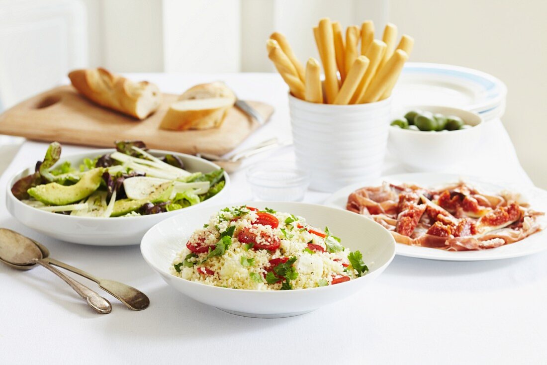 Mediterranes Buffet mit Parmaschinken, Avocado-Fenchel-Salat, Oliven und Gemüse-Couscous, dazu Brot und Grissini
