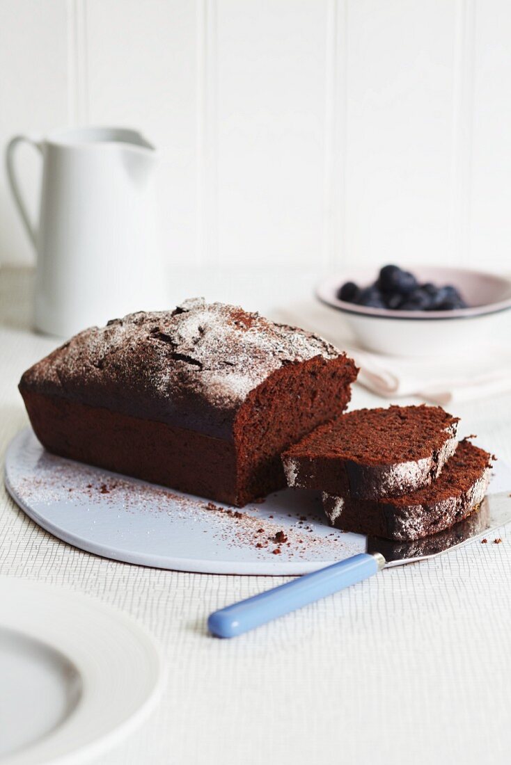 A chocolate loaf cake dusted with icing sugar with a bowl of blueberries in the background