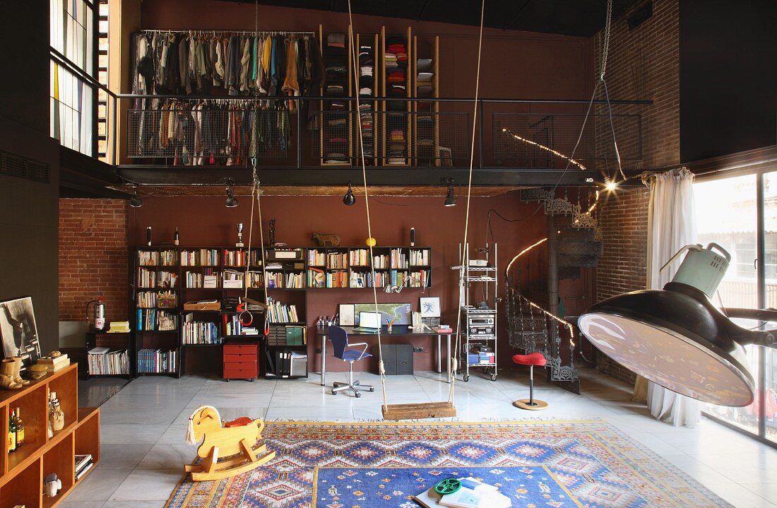 Double-height loft interior with bookcase and desk below open-fronted wardrobes on mezzanine; swing in foreground next to rocking horse on large rug