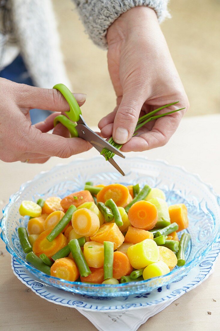 A woman cutting chives into a bowl of carrots and beans