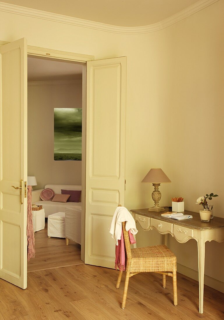 Rattanstuhl vor zierlichem Wandtisch mit Schnitzereien, neben offener Flügeltür und Blick ins Wohnzimmer