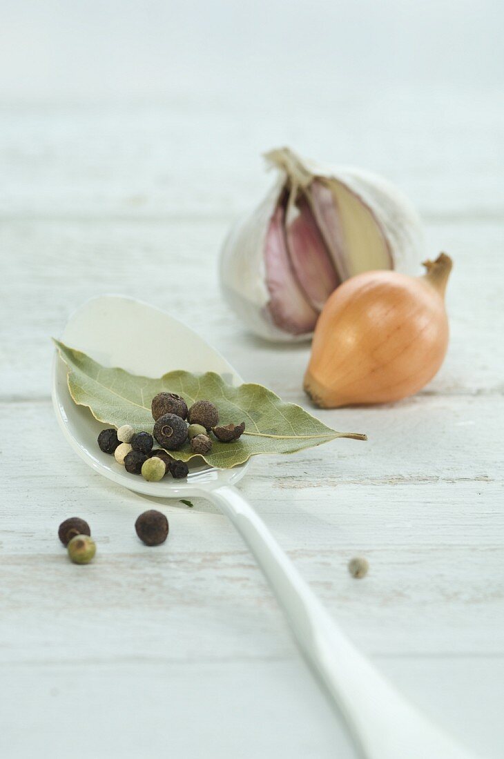 A bay leaf, pepper corns and allspice berries on a spoon with an onion and a bulb of garlic in the background
