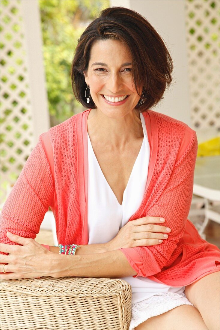 A dark-haired woman sitting in a wicker chair wearing a top and a cardigan
