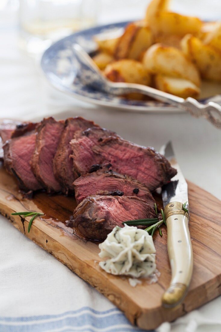 Aufgeschnittenes Rindersteak auf Schneidebrett mit Butter, dahinter Bratkartoffeln