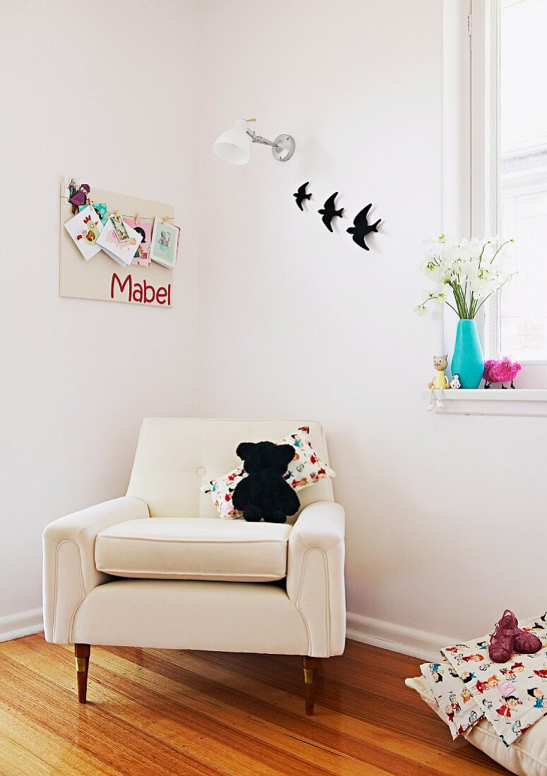 Soft toy sitting on elegant, white armchair in corner of child's bedroom below pinboard and bird ornaments on wall