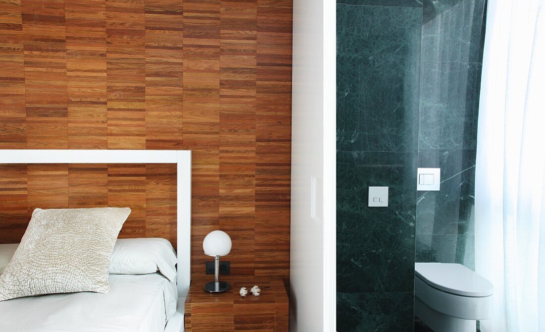 Double bed with frame-like headboard and ensuite bathroom with polished stone tiles; wall and bedside table made from parquet-style wood panelling